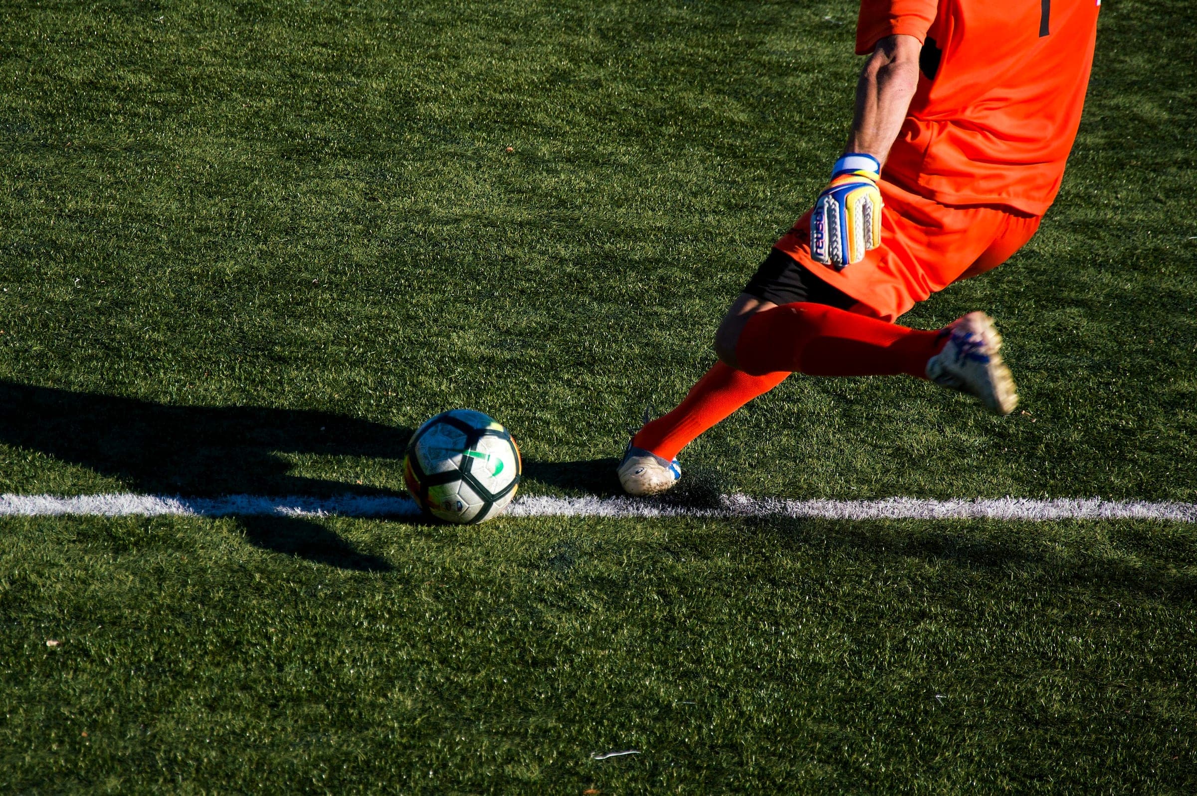 Goalkeeper in orange striking a football off the line on the grass